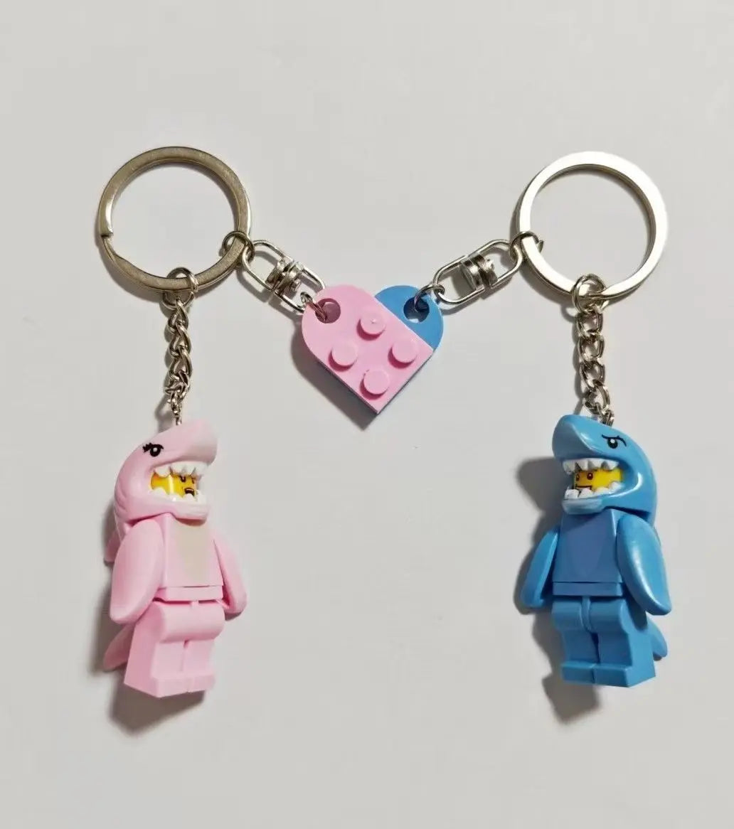 A keychain set featuring two shark-themed building block charms, one pink and one blue, attached to silver split rings.