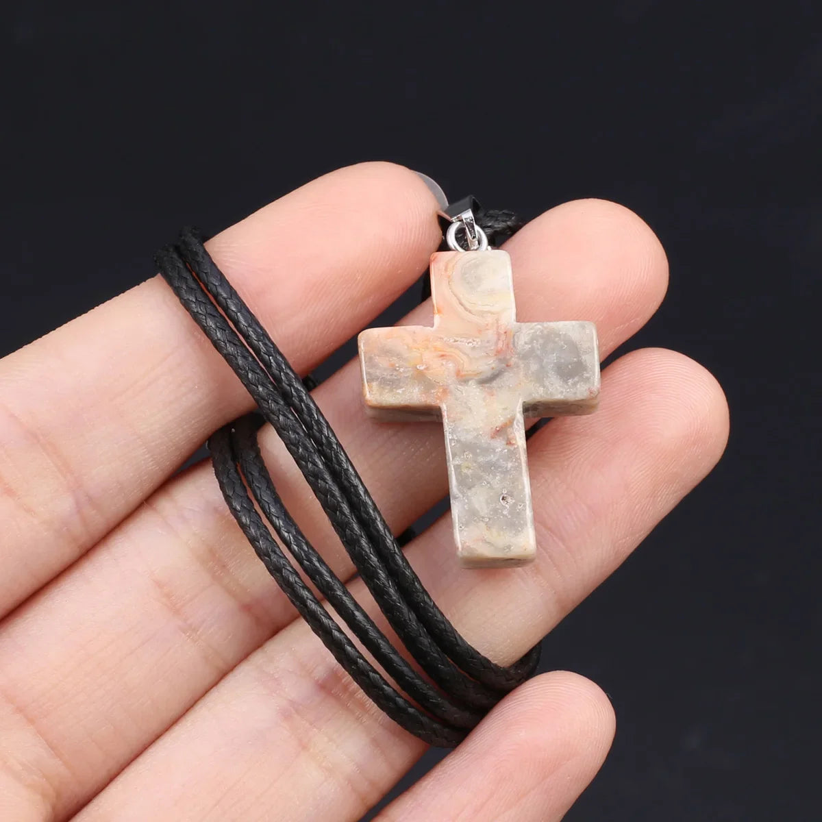 Eine Hand hält eine Tigerauge-Achat-Kreuz-Anhängerkette mit schwarzem Band vor dunklem Hintergrund.