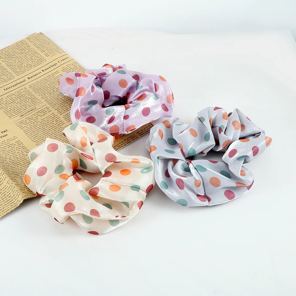 A set of three colorful knitted hair scrunchie headbands in shades of purple, pink, and gray are displayed on a light backgro