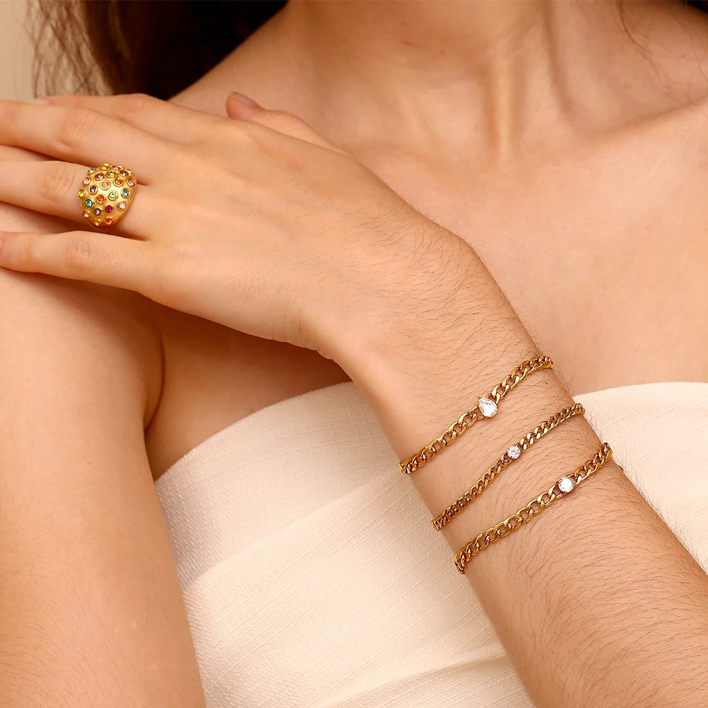 A close-up of a hand wearing a gold charm bracelet with small white stones and a gold ring.