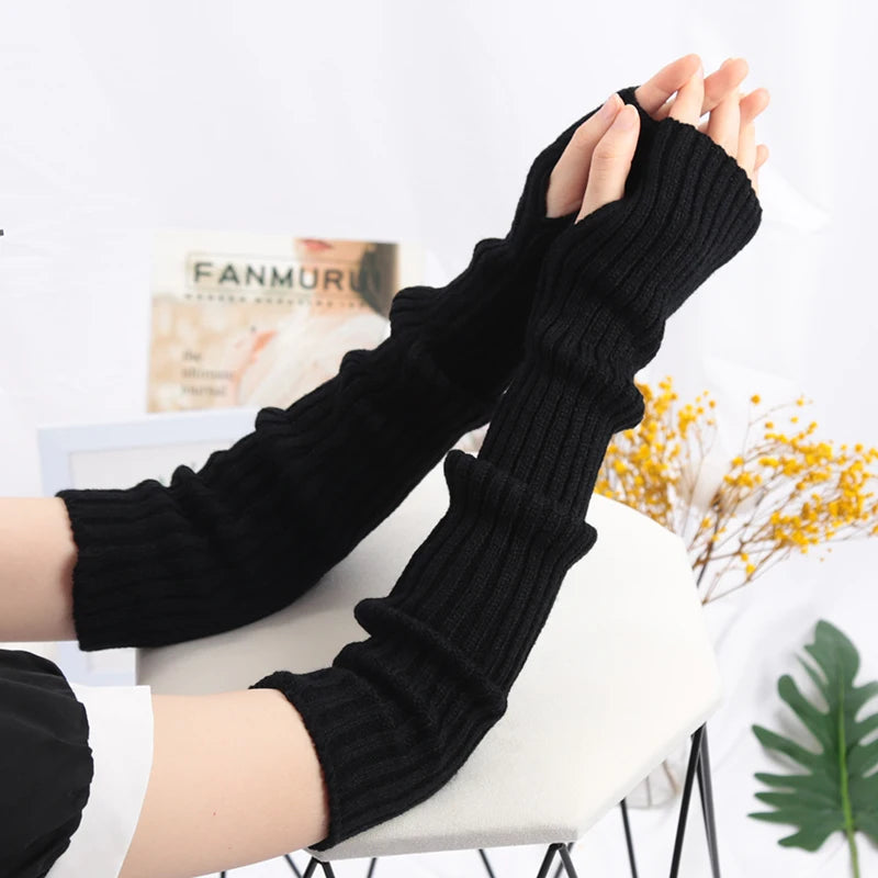 A close-up of black knitted fingerless gloves being worn on a hand, displayed against a white background.