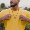 Un hombre con una camiseta amarilla muestra un collar de cruz de acero inoxidable negro.