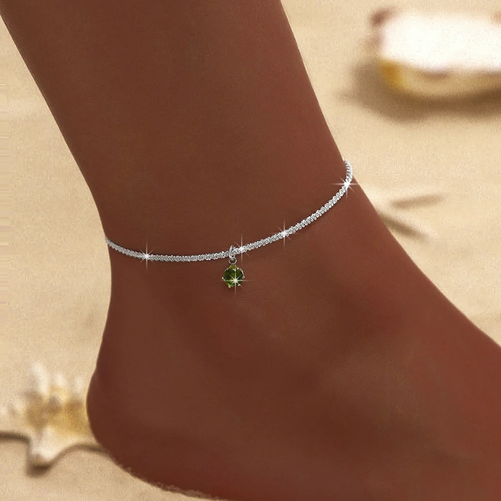 A close-up of a delicate silver plated CZ anklet with a green gemstone dangling on a person's ankle.