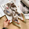 A silk scarf tied in a bow with horse motifs is displayed on a white paper next to a hand.