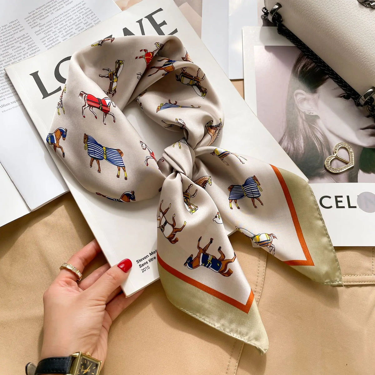 A silk scarf tied in a bow with horse motifs is displayed on a white paper next to a hand.