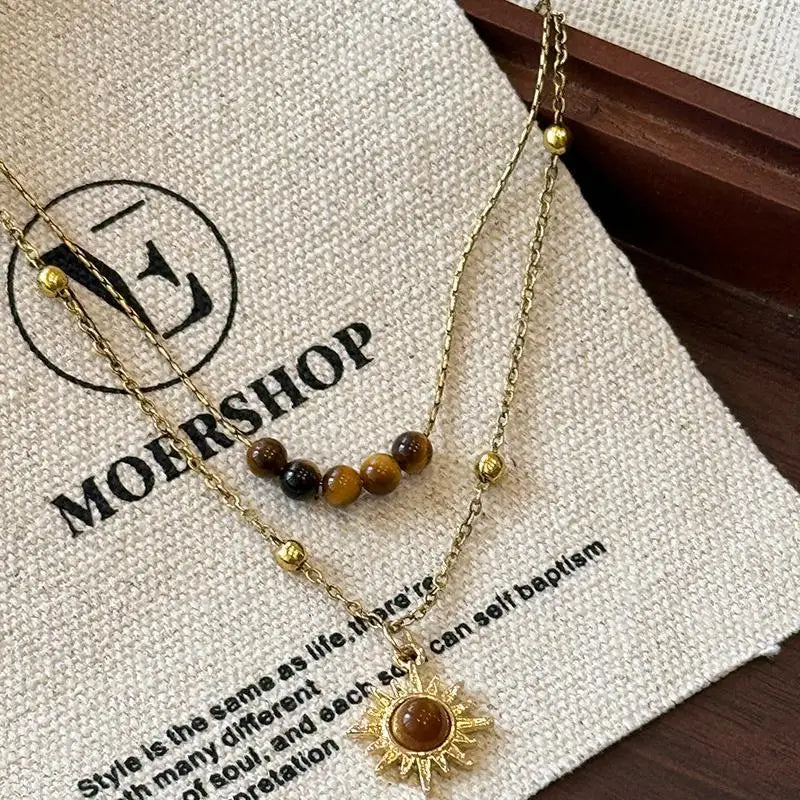 Gold chain necklace with a tiger eye sunflower pendant displayed on a beige background.