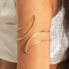 A close-up of a person's arm showing three gold-plated swirl cuff armbands worn on the wrist.