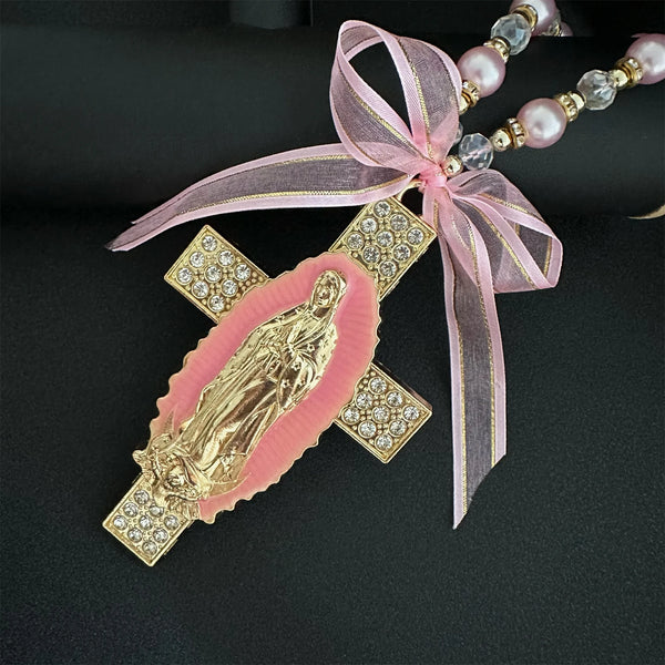 A gold-colored religious charm featuring the Virgin Mary, set within a cross design and tied with a pink ribbon.