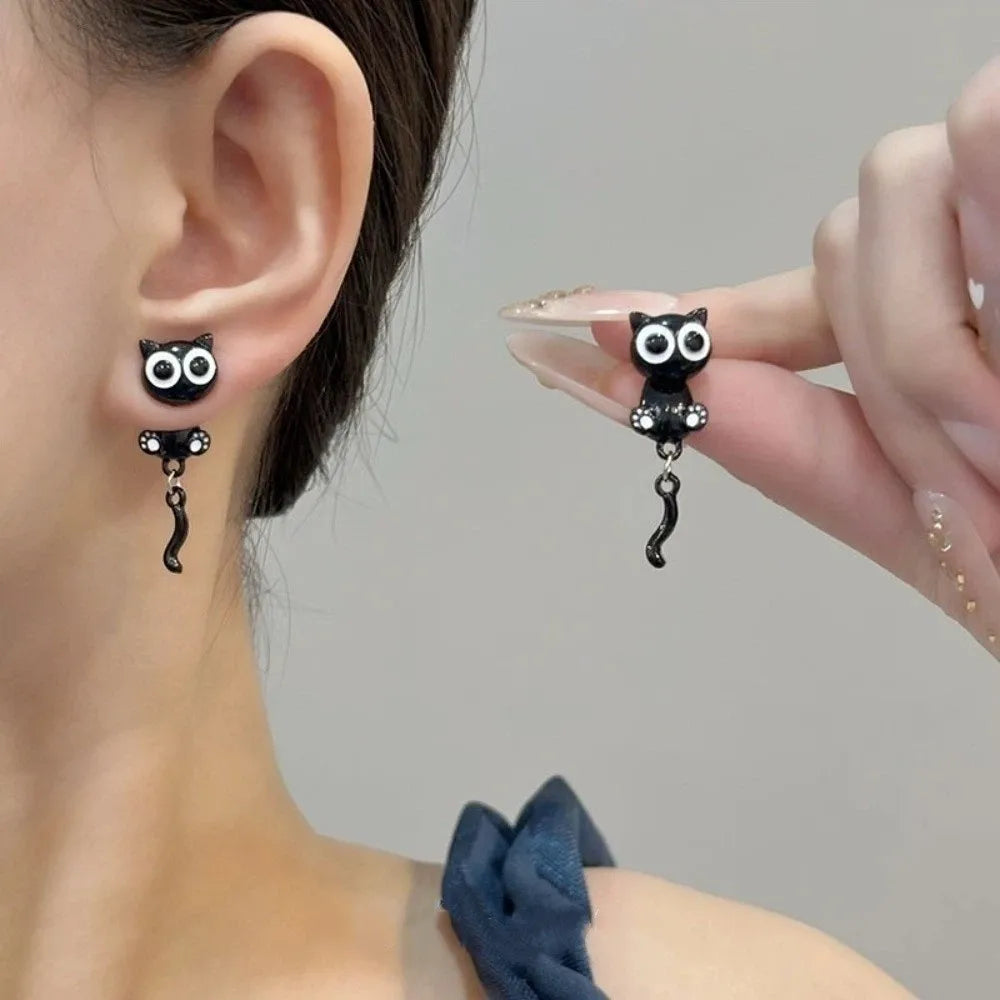 A close-up of black cat stud earrings worn on a woman's ear, held by a hand.