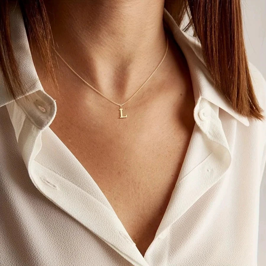 A close-up of a woman wearing a delicate gold initial letter necklace with a small L charm against a white collared shirt.