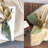 A silk scarf with a floral print in muted green and beige tones is displayed on a light background.