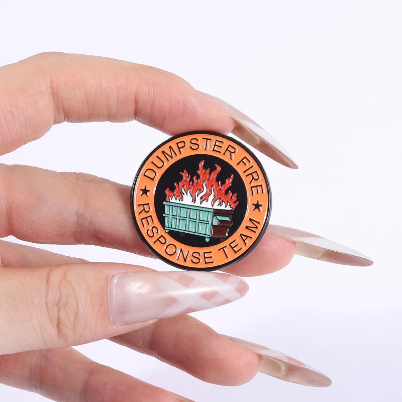 Eine Hand hält eine schwarze runde Anstecknadel mit einem stilisierten orangefarbenen Müllbranddesign und dem Text DUMPSTER F