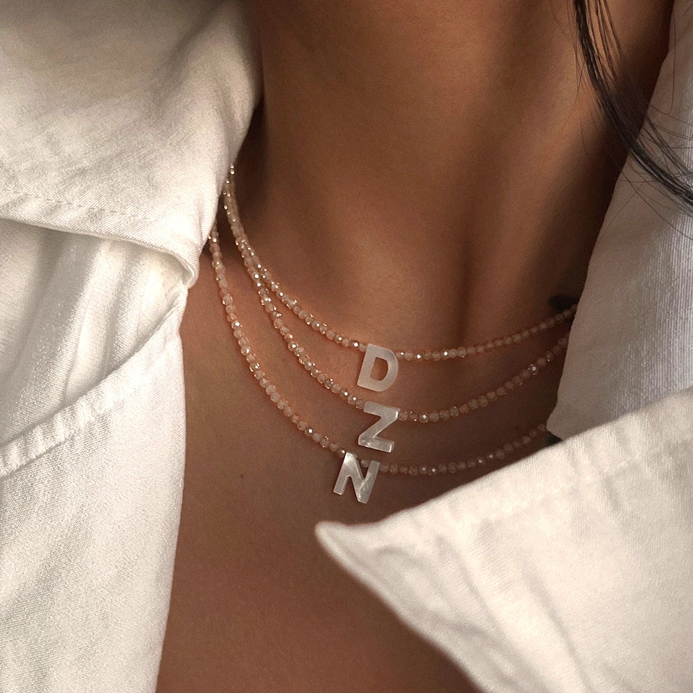 A close-up of a delicate rose gold crystal necklace featuring the initial DNZ charm worn against a white shirt.