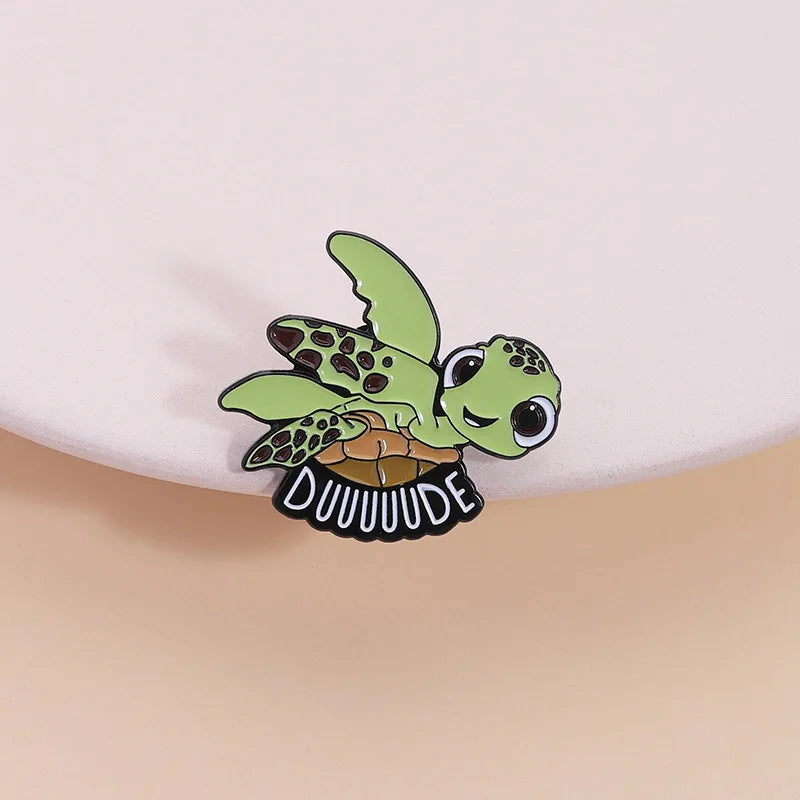 A cute sea turtle enamel lapel pin brooch featuring green and brown scales with the name DUUUUUDE engraved below.