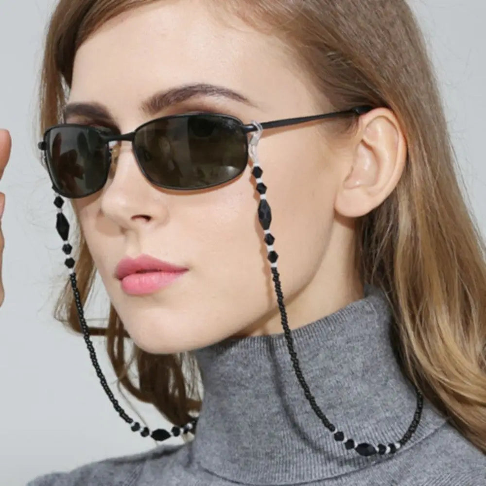 A close-up of a woman wearing dark sunglasses with a pearl lanyard and chain against a light background.