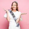 A young woman wearing a white dress and a silver tiara crown sash for girls, smiling and gesturing with her hands.