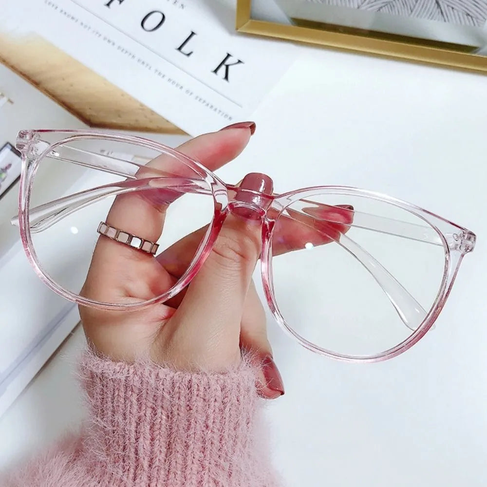A hand holds a pair of pink, clear-framed anti-blue light spectacles against a white background.
