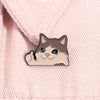 A cute enamel brooch pin of a cat with sad eyes and a smiling mouth on pink fabric.