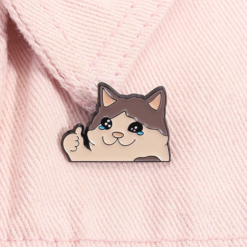 A cute enamel brooch pin of a cat with sad eyes and a smiling mouth on pink fabric.