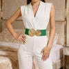 A woman models a white suit with a green and gold elastic waist belt against a luxurious bedroom background.