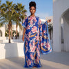 Une femme modélise une longue robe maxi florale bleue dans un cadre extérieur lumineux.