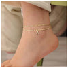 A close-up of a woman's ankle showing layered gold letter anklets with an 'A' charm.
