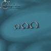 Three silver titanium steel nose rings with small crystal accents displayed against a teal background.