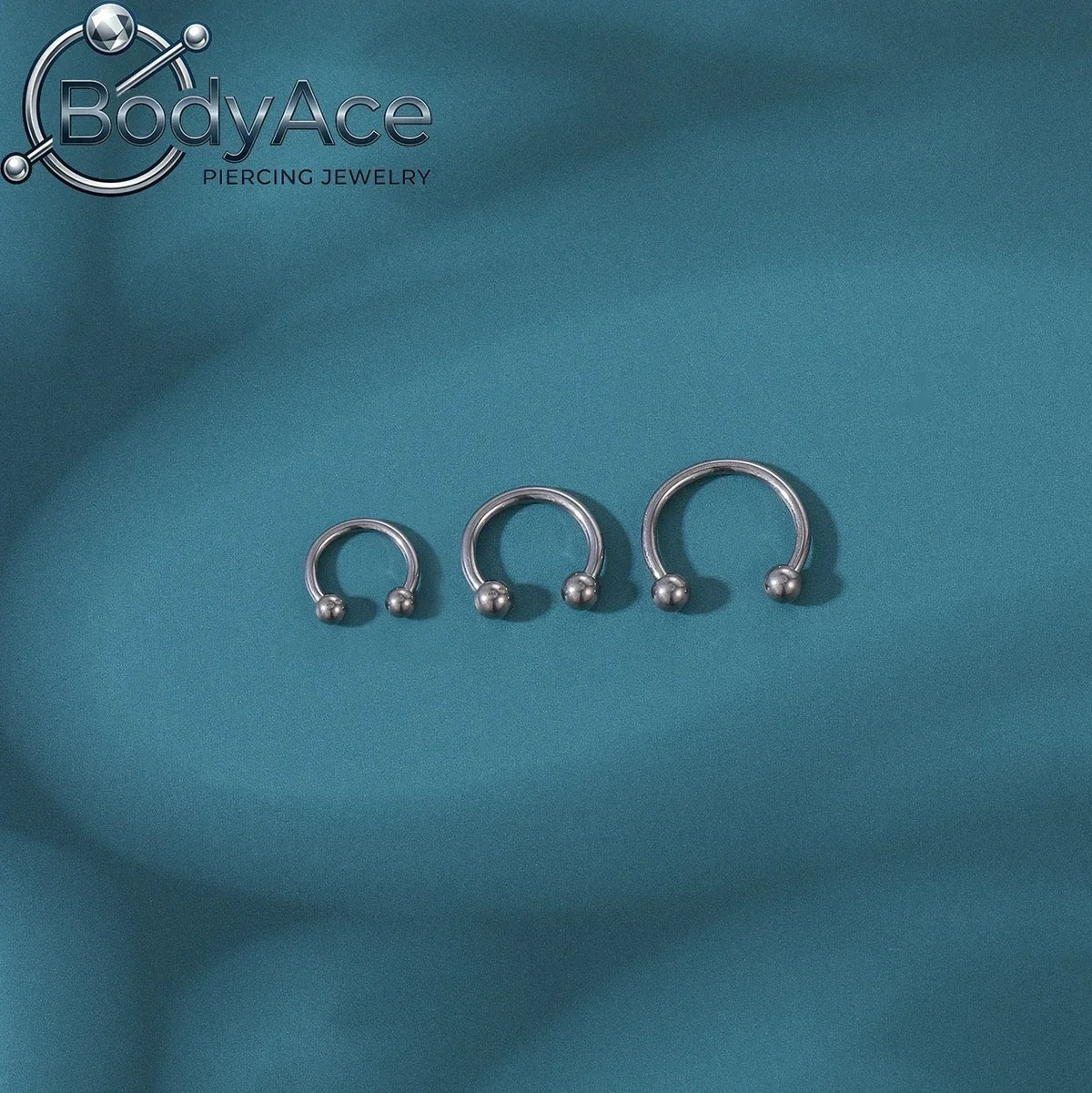 Three silver titanium steel nose rings with small crystal accents displayed against a teal background.