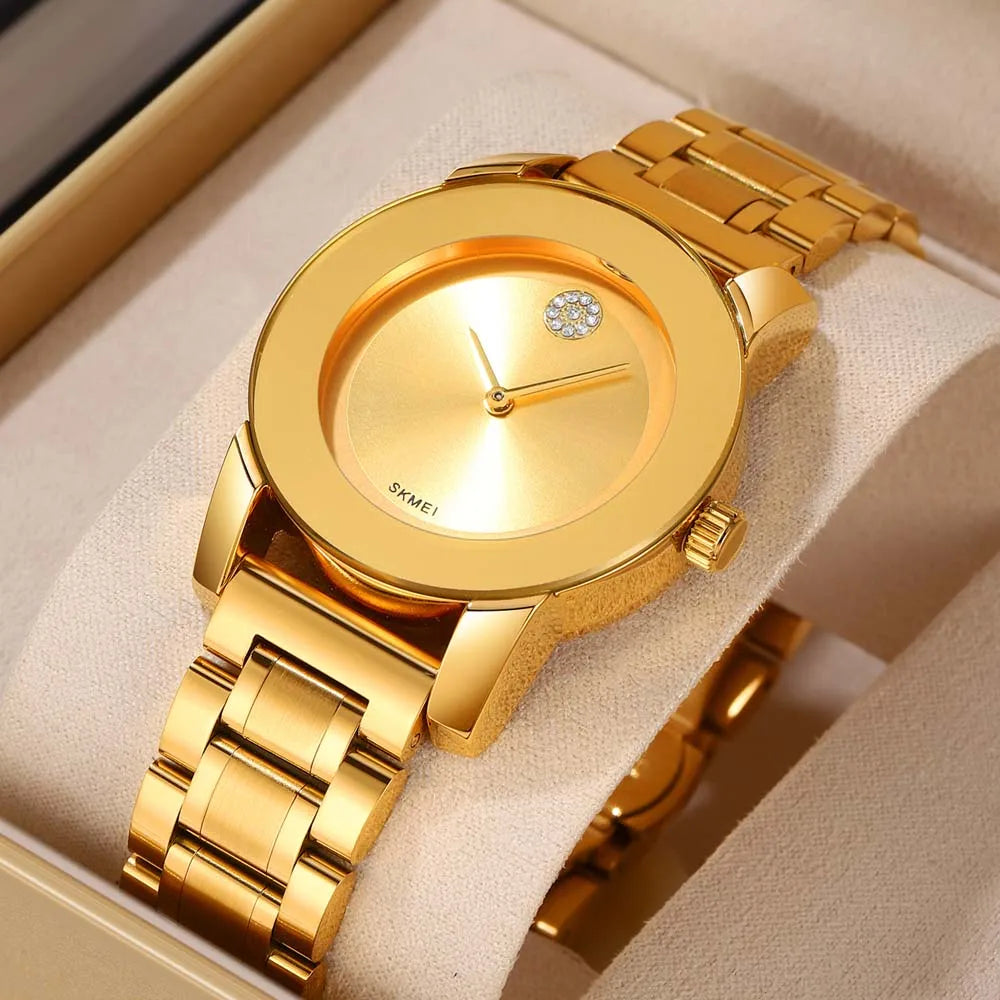 A gold-toned minimalist quartz wristwatch with a round face and a textured metal band is displayed in a light-colored box.