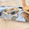A gold-toned quartz watch with a round face and Roman numerals is displayed on a silver tray next to a piece of driftwood.