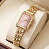 Reloj de pulsera analógico de cuarzo tono dorado con esfera rosa y bisel cuadrado, mostrado en una caja blanca.