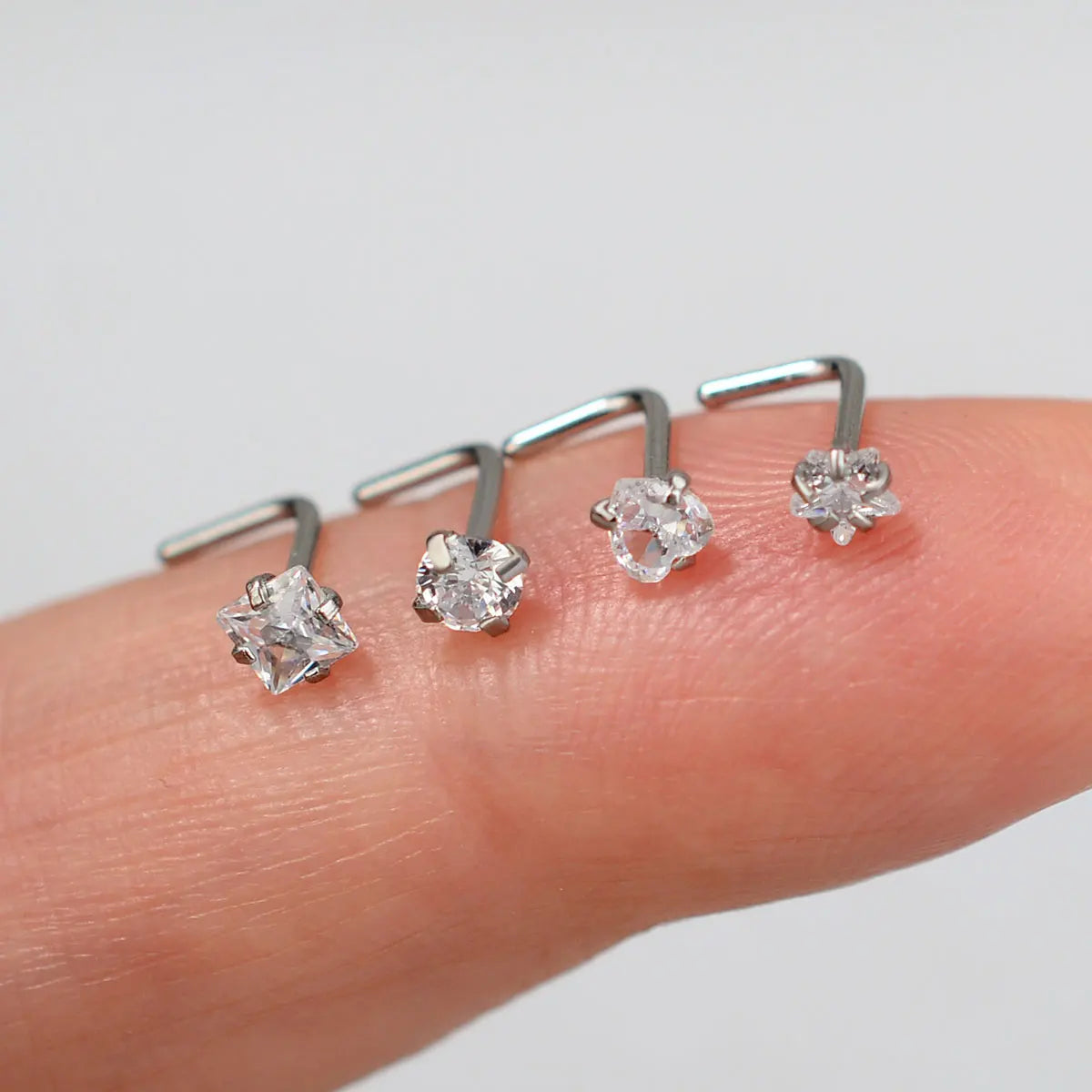 Una mano sostiene varios pendientes en forma de L de acero inoxidable con piedras preciosas transparentes.