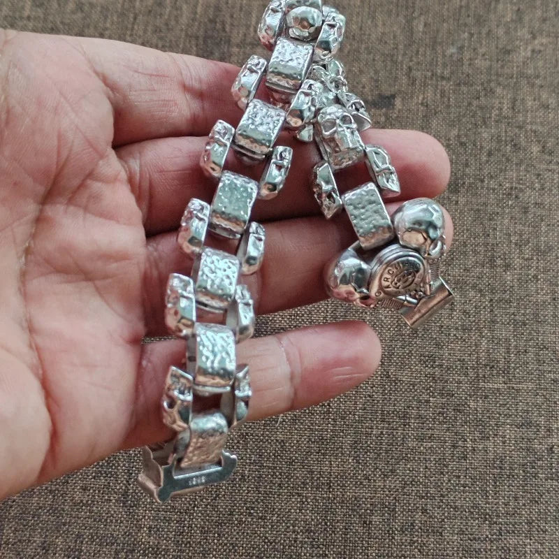 Eine Hand hält ein silbernes Totenkopf-Armband aus Vintage-Stil für Männer mit rechteckigen und Totenkopf-Charms.