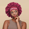 A woman wearing a deep magenta satin sleep bonnet with a matching wrap band.