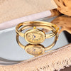 A gold-toned elegant quartz watch is displayed on a reflective surface next to a wooden anatomical model.