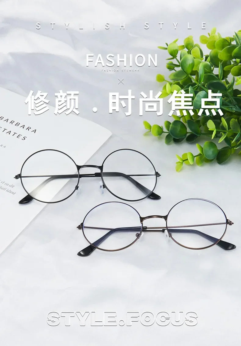 Un par de gafas redondas de metal con monturas finas sobre un fondo blanco junto a follaje verde.