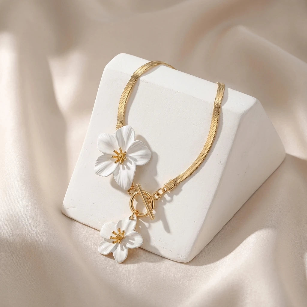 A delicate gold chain necklace with white flower pendants rests on a white geometric block against a soft beige fabric backgr