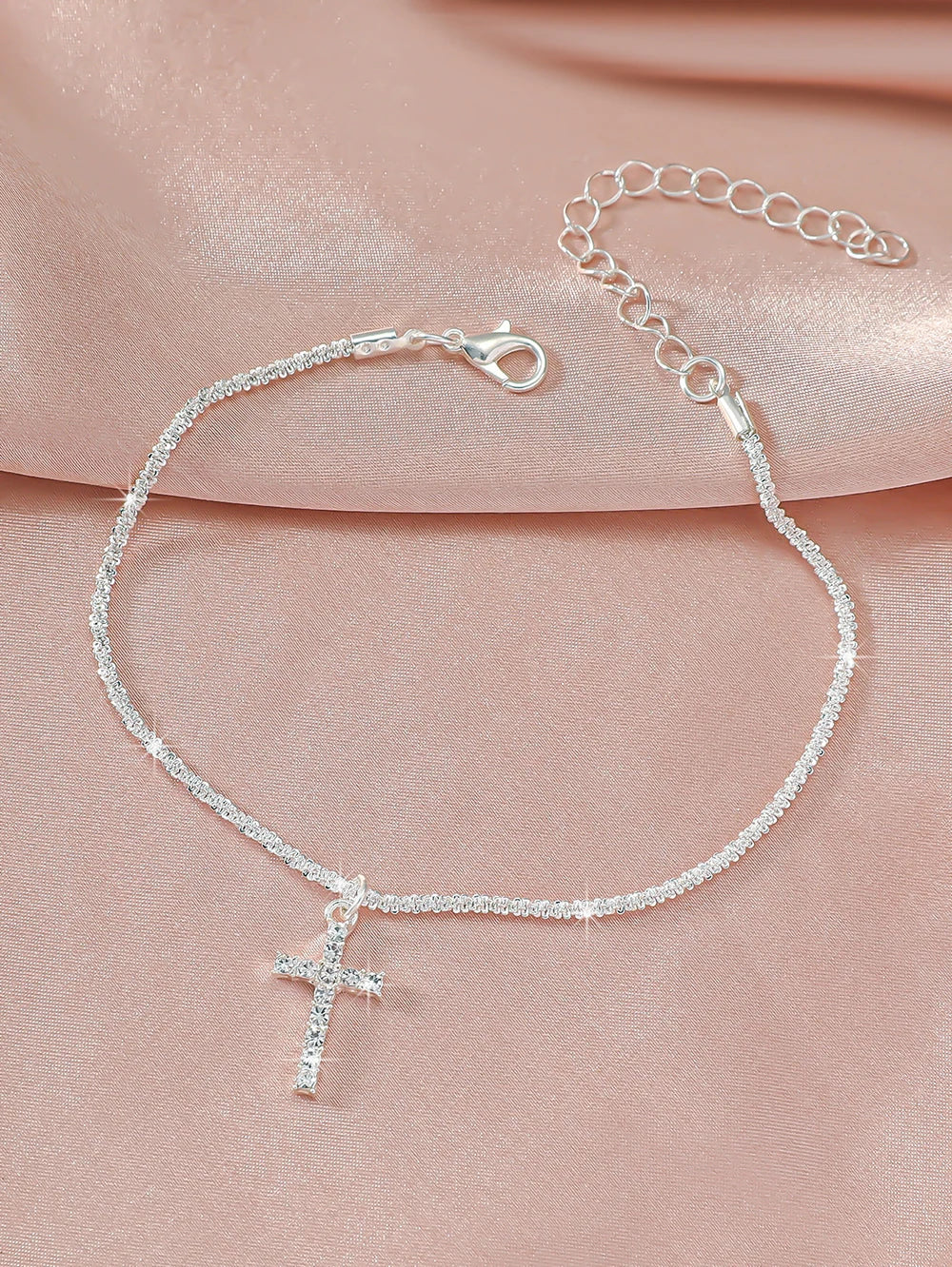 A delicate sparkling zircon heart cross anklet is displayed against a soft pink fabric background.