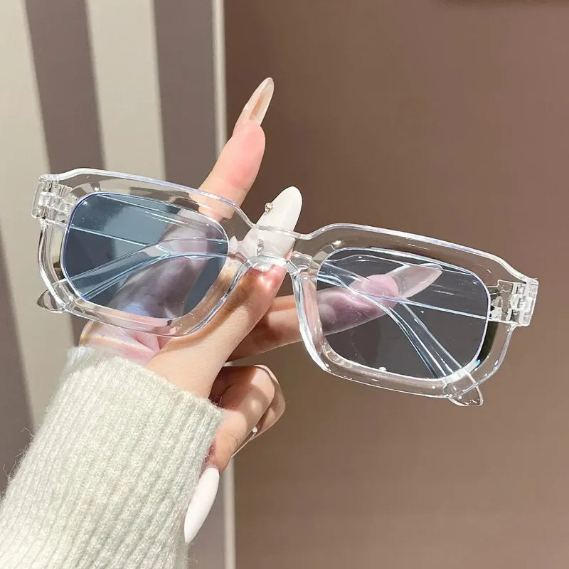 A hand holds a pair of clear plastic retro square fashion sunglasses with blue lenses against a neutral background.