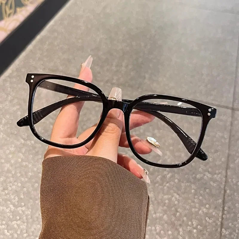 A hand holds a pair of black, classic square anti-blue light eyeglasses against a light gray background.