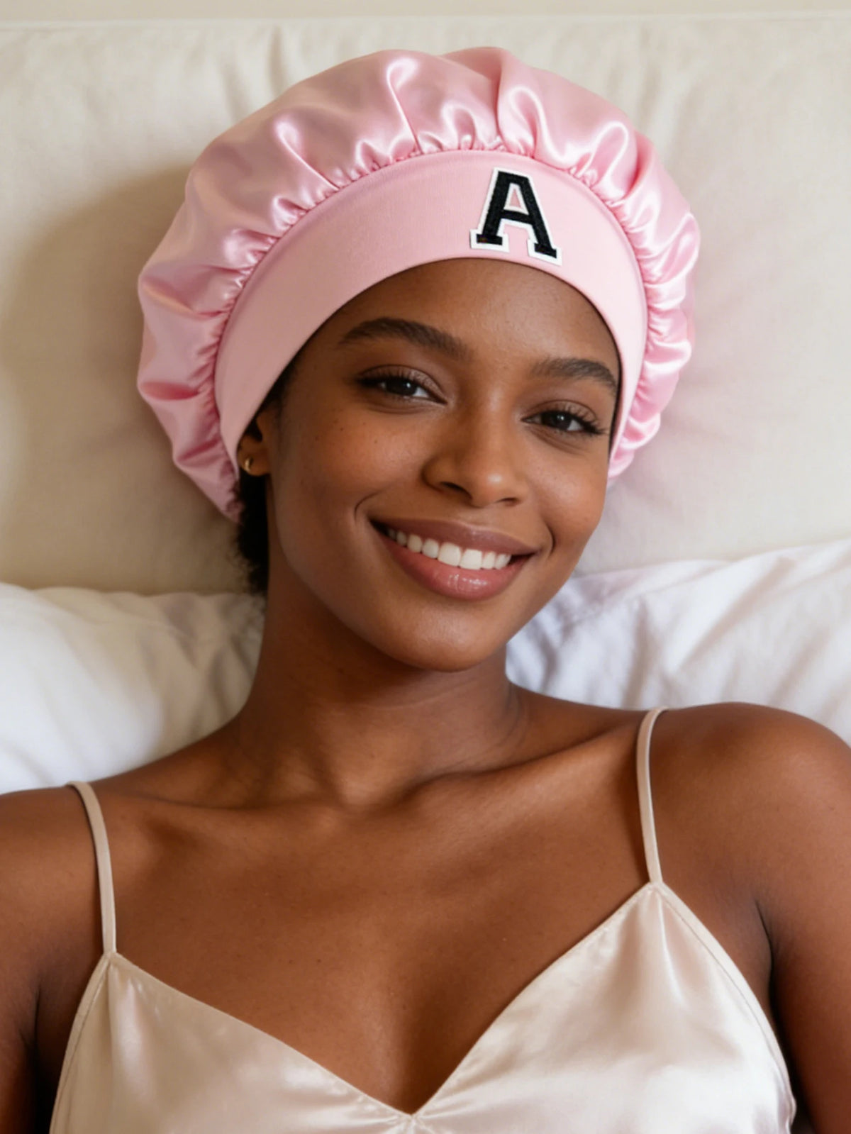 A smiling woman wearing a pink satin hair cap with a letter A on the front, lying in bed.