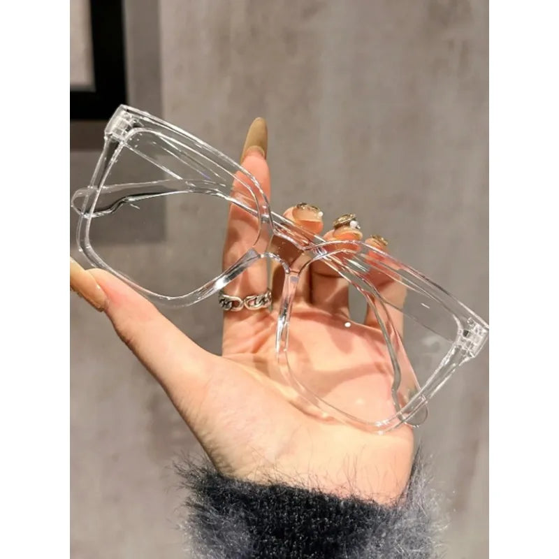 Una mano sostiene unas gafas cuadradas transparentes con detalles plateados contra un fondo desenfocado.