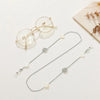 A delicate silver chain necklace featuring pearl and clear gemstone charms, displayed next to a pair of round metal-framed ey