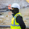 A person wearing a white hard hat, clear goggles, and a black balaclava and high-visibility vest is visible outdoors.